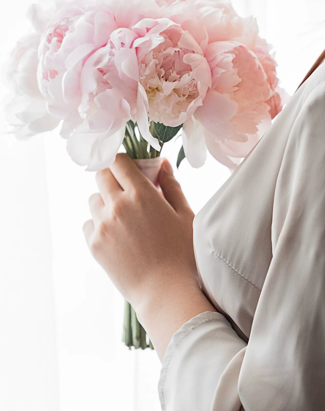 A bride's hand holds a bouquet of blush peonies by a bright window.